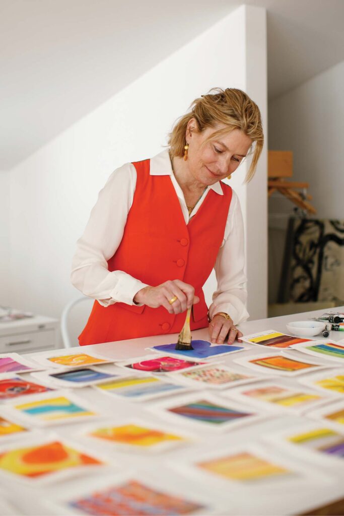 Isa Catto wearing a red vest over a white long sleeve shirt. She paints in blue, surrounded by her other paintings, varying in different colors.